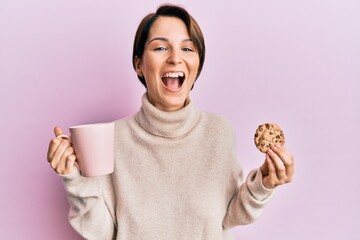 Young brunette woman with short hair drinking a cup of coffee and cookie smiling and laughing hard out loud because funny crazy joke.