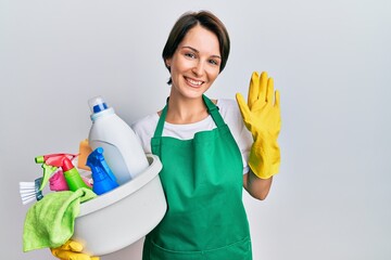 Young brunette woman with short hair wearing apron holding cleaning products waiving saying hello happy and smiling, friendly welcome gesture © Krakenimages.com