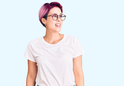 Young beautiful woman with pink hair wearing casual clothes and glasses looking away to side with smile on face, natural expression. laughing confident.