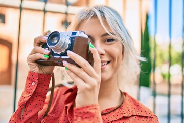 Young blonde woman smiling happy using vintage camera walking at the city.