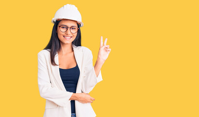 Fototapeta na wymiar Young beautiful latin girl wearing architect hardhat and glasses smiling with happy face winking at the camera doing victory sign. number two.