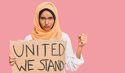 Young brunette arab woman holding united we stand banner annoyed and frustrated shouting with...