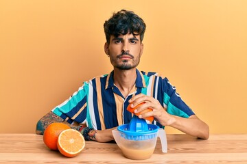 Young hispanic man sitting on the table using juicer thinking attitude and sober expression looking...