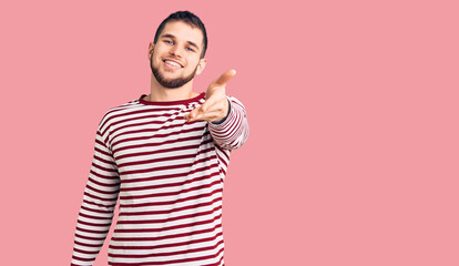Young handsome man wearing striped sweater smiling cheerful offering palm hand giving assistance and acceptance.