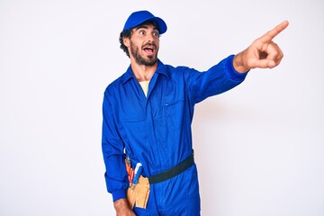 Handsome young man with curly hair and bear weaing handyman uniform pointing with finger surprised ahead, open mouth amazed expression, something on the front