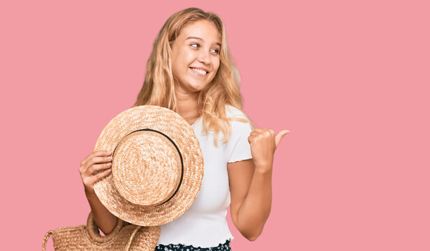 Young blonde girl holding summer hat and wicker handbag pointing thumb up to the side smiling happy with open mouth