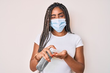 Young african american woman with braids wearing medical mask using hand sanitizer winking looking...