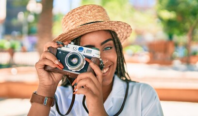 Young african american woman with braids smiling happy taking pictures with vintage camera outdoors on a sunny day of summer