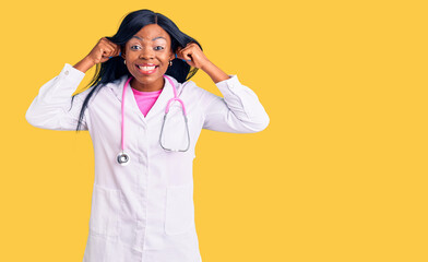 Young african american woman wearing doctor stethoscope smiling pulling ears with fingers, funny...