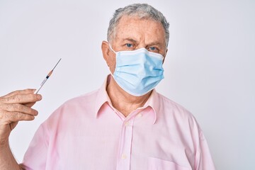 Senior grey-haired man wearing medical mask holding syringe thinking attitude and sober expression looking self confident