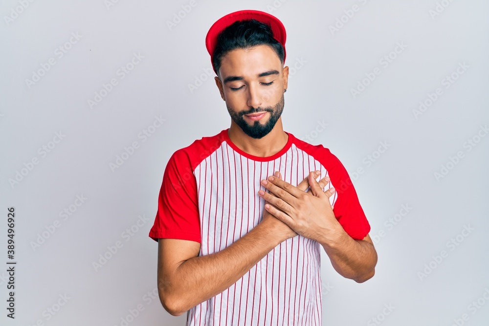 Wall mural Young man with beard wearing baseball uniform smiling with hands on chest, eyes closed with grateful gesture on face. health concept.