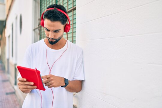 Young arab man with serious expression using headphones and touchpad at the city.