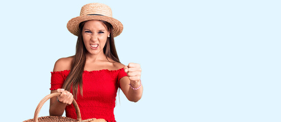 Young beautiful hispanic woman wearing summer hat and holding picnic wicker basket with bread...