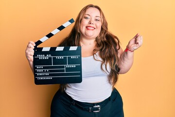 Plus size caucasian young woman holding video film clapboard screaming proud, celebrating victory...
