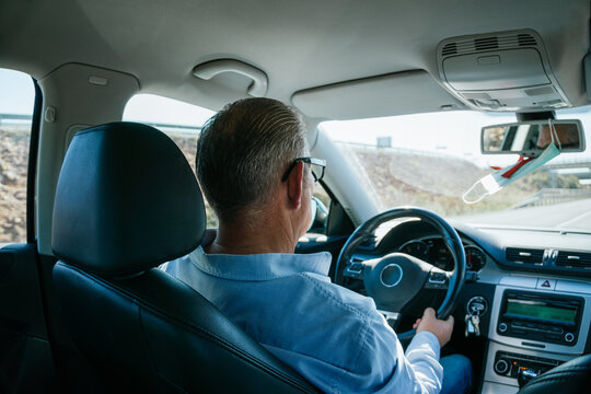 Caucasian male with gray hair and about 50 years old driving a car on the road. The driver is dressed in a blue shirt and wears eye glasses. The car is spacious and stately.