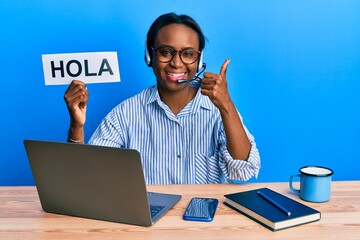 Young african woman wearing operator headset holding hola word smiling happy and positive, thumb up doing excellent and approval sign © Krakenimages.com