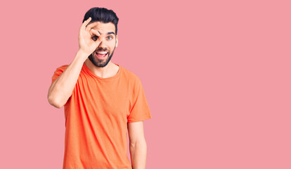 Young handsome man with beard wearing casual t-shirt doing ok gesture with hand smiling, eye looking through fingers with happy face.