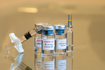 Closeup view of a set of three COVID-19 vaccination drug dose trial vials with a filled injection syringe in the front with a tiny droplet escaping from the needle tip - 025