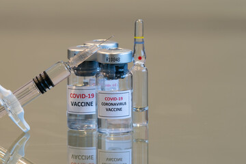 A closeup set of COVID-19 vaccination drug dose trial vials with a filled injection syringe in the front with a tiny droplet escaping from the needle tip with copy space - 048