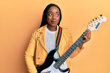 Young african american woman playing electric guitar smiling looking to the side and staring away...
