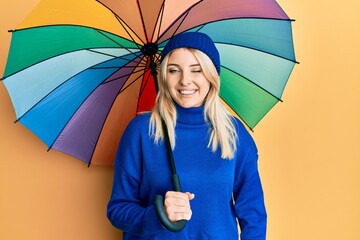 Young caucasian woman holding colorful umbrella winking looking at the camera with sexy expression,...
