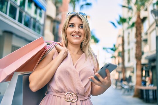 Young blonde woman smiling happy holding shopping bags and using smartphone at the city.