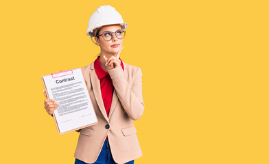 Young beautiful woman wearing architect hardhat holding clipboard with contract document serious...