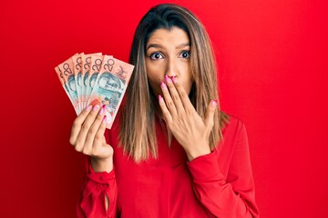 Beautiful brunette woman holding australian 20 dollars covering mouth with hand, shocked and afraid for mistake. surprised expression