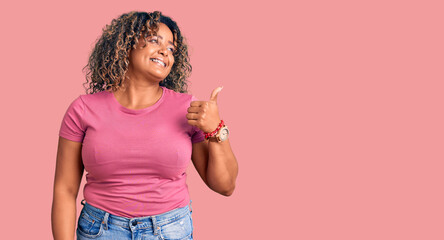 Young african american plus size woman wearing casual clothes smiling with happy face looking and pointing to the side with thumb up.