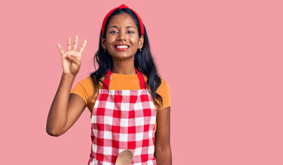 Young indian girl wearing professional baker apron showing and pointing up with fingers number four...