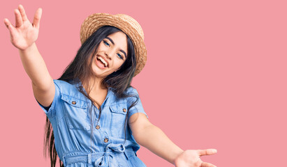 Brunette teenager girl wearing summer hat looking at the camera smiling with open arms for hug. cheerful expression embracing happiness.
