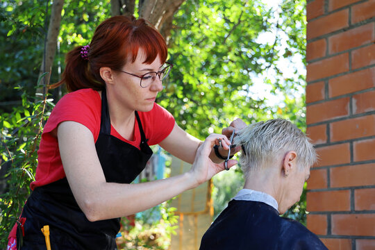 Young red-haired woman hairdresser makes short haircut to woman outdoors. She is focused on her work. Real not staged scene