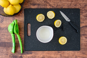 Fresh lemon juice in a white bowl, lemon halves on a black cutting board, paring knife, wood table,...
