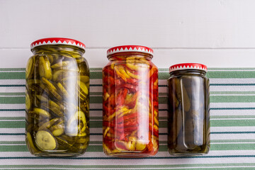 jars with pickled cucumbers and pepper on striped fabric white wooden surface background.