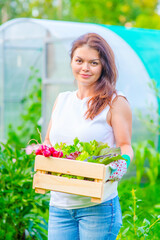 A young woman farmer stands on a greenhouse in the garden and holding a box with the harvested greens and vegetables © Ermolaeva Olga