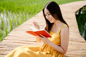 Asian traveler woman in yellow dress reading red book on wooden bridge across rice field.