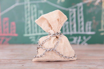 A money bag chained in front of a blackboard with financial charts