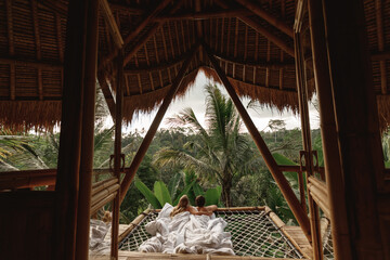 Fototapeta na wymiar Young caucasian travel couple lie in hammock in bamboo eco house, nature and mountains on background. Morning in tropical resort on Bai island