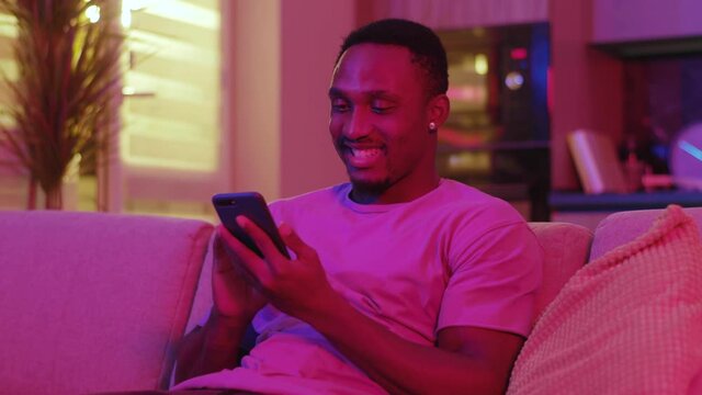 Joyful african american stylish happy man receiving messages in smartphone browsing funny video application staying home in bright colored neon illumination.