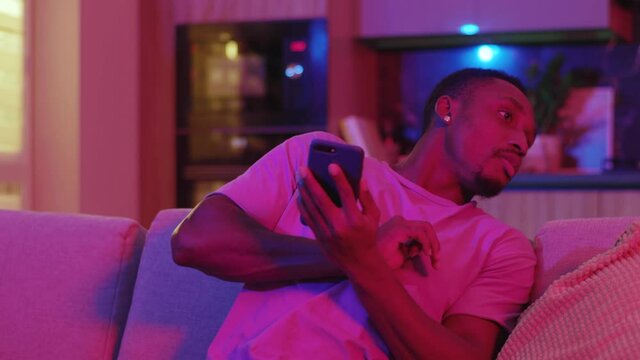 Tricky funny african american guy using smartphone watching TV hiding his cellphone under sofa laughing when girlfriend comes in. Nomophobia. Emotions.