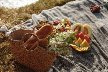 Beautiful summer picnic in boho style. Organic fresh fruits, wicker bag with baguettes and flowers on linen blanket. Picnic background with basket, fruits and wicker bag with baguette