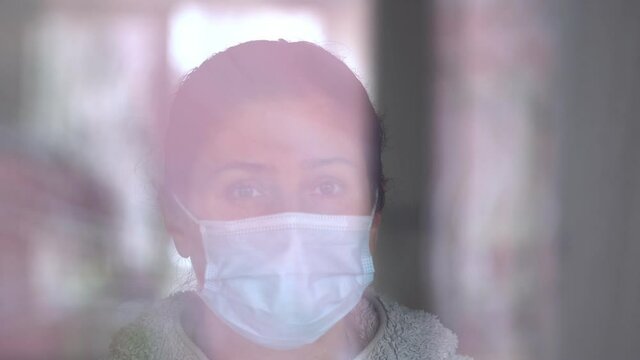 Young caucasian woman in a coronavirus mask touching with her hand to the glass of the window. Sad woman in quarantine. Pensive young woman in mask to protect herself from covid-19 while looking out