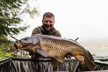 Happy smiling fisherman holding his fish  with lake on the background. Selective focus on the carp.