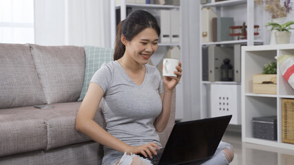 cheerful female blogger in casual wear drinking cup of coffee and uploading new articles on her page while sitting on living room floor and using laptop.