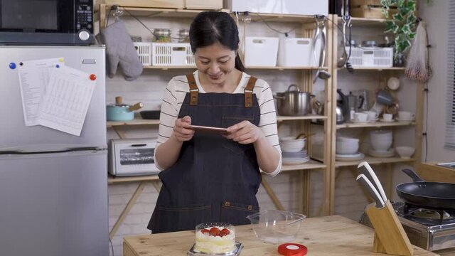 cheerful asian girl running a small home baking business by kitchen table, using phone to take pictures for her product from different angles.