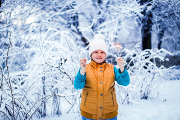 Fototapeta premium Kid girl in yellow winter vest and white knitted hat on a snow scooter with sparklers with the first snow, snowfall and a walk in the park in the backyard. Frost and snowy weather.