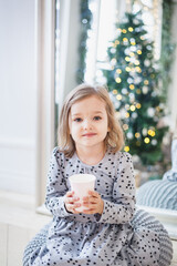 The room is decorated for the new year, the girl is sitting near the mirror, holding a glass or mug with a drink, milk or juice, Christmas