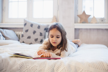 The room is decorated for the new year, the baby girl is lying on the bed and reading a book, learning, fairy tales, fun and childhood