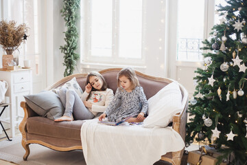 The room is decorated for the new year, two girls on the sofa with a book, puzzles, Christmas morning, communication of children, sisters