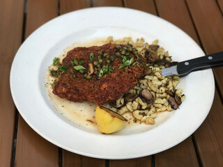 Fried chicken schnitzel with lemon and spatzle pasta with mushrooms is shown on a plate. Additionally, this updated version of the German classic is served with capers and green onions on the chicken.